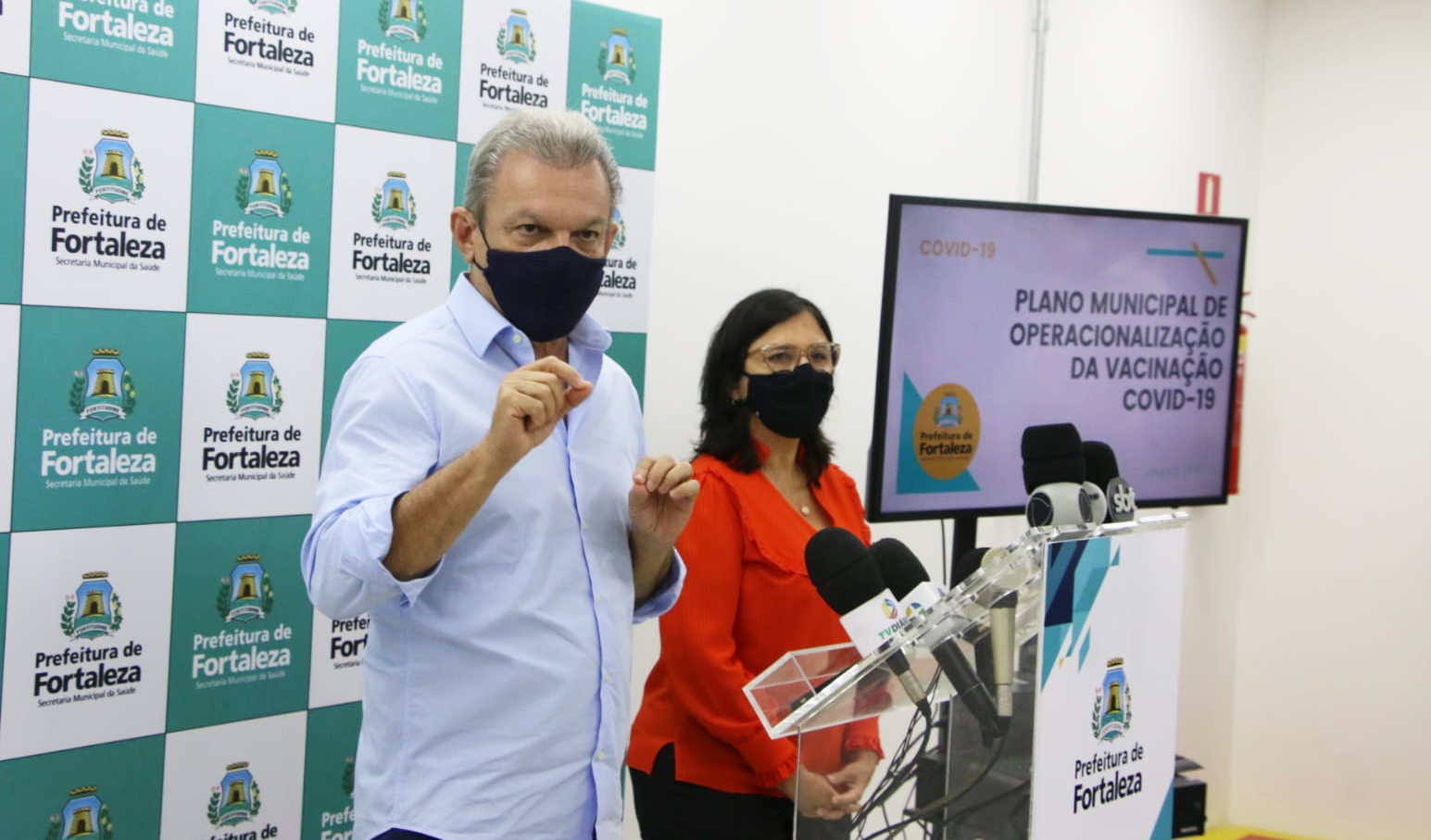 José Sarto ao lado da secretária Ana Estela gesticulando atrás de púlpito e monitor ao lado da secretária onde está escrito Plano Municipal de Operacionalização da Vacinação Covid-19